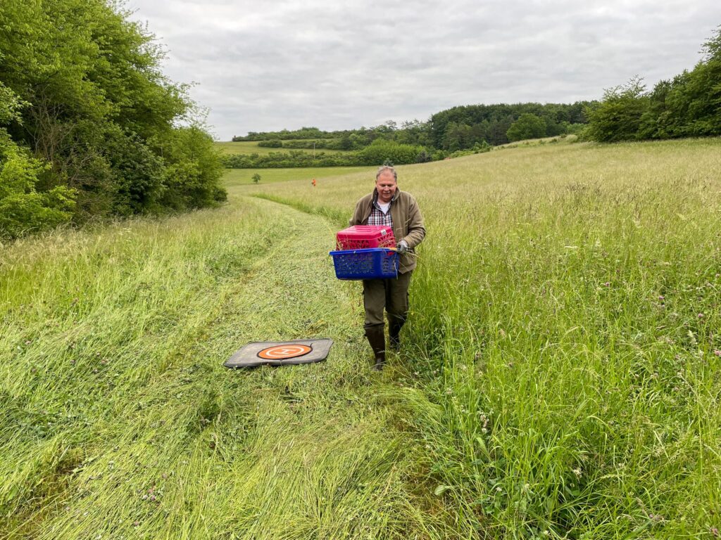 Joerg Hartmann trägt ein gerettetes Rehkitz aus der Wiese. Rechts neben ihm sieht man den Landeplatz für die Drohne. Foto: Jutta Adam-Fuß